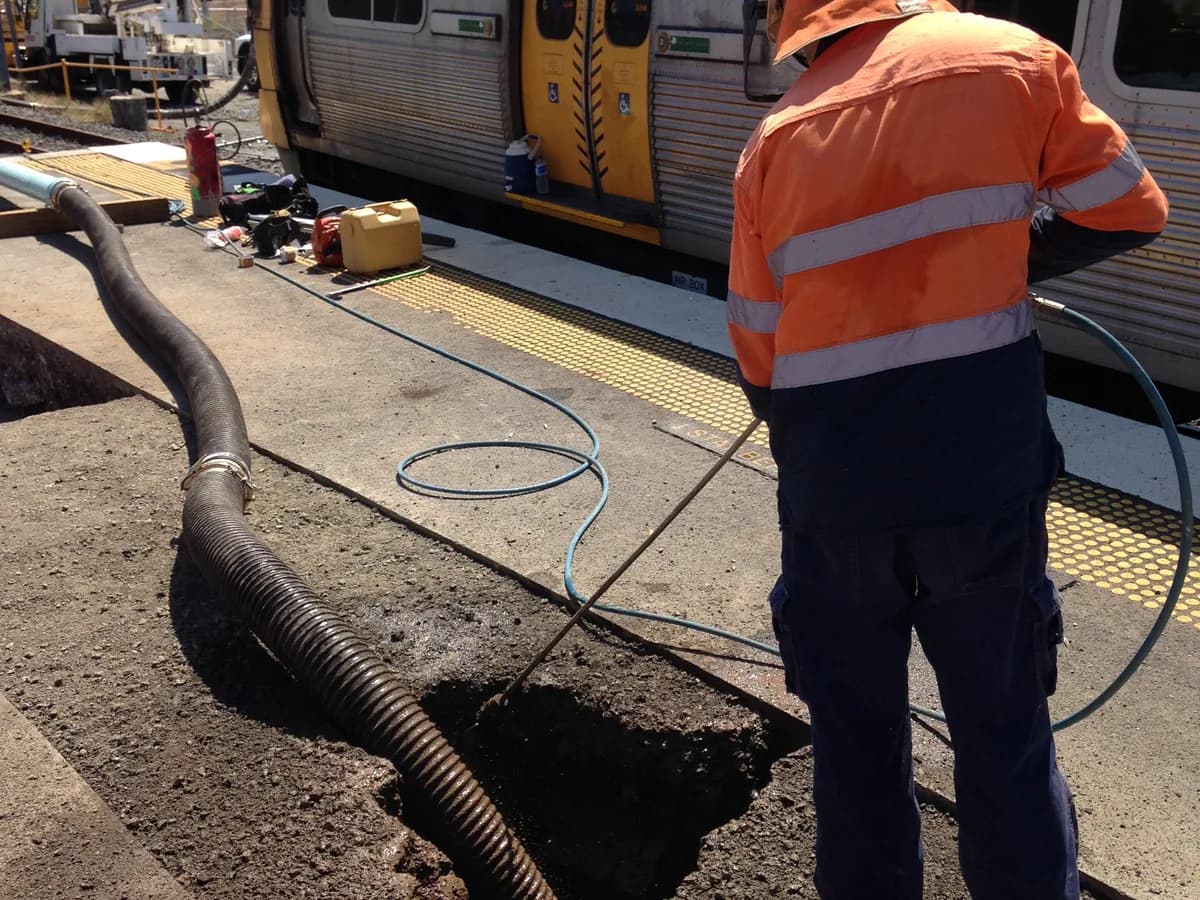 Worker using a water lance beside a vacuum hose during potholing work near rail infrastructure